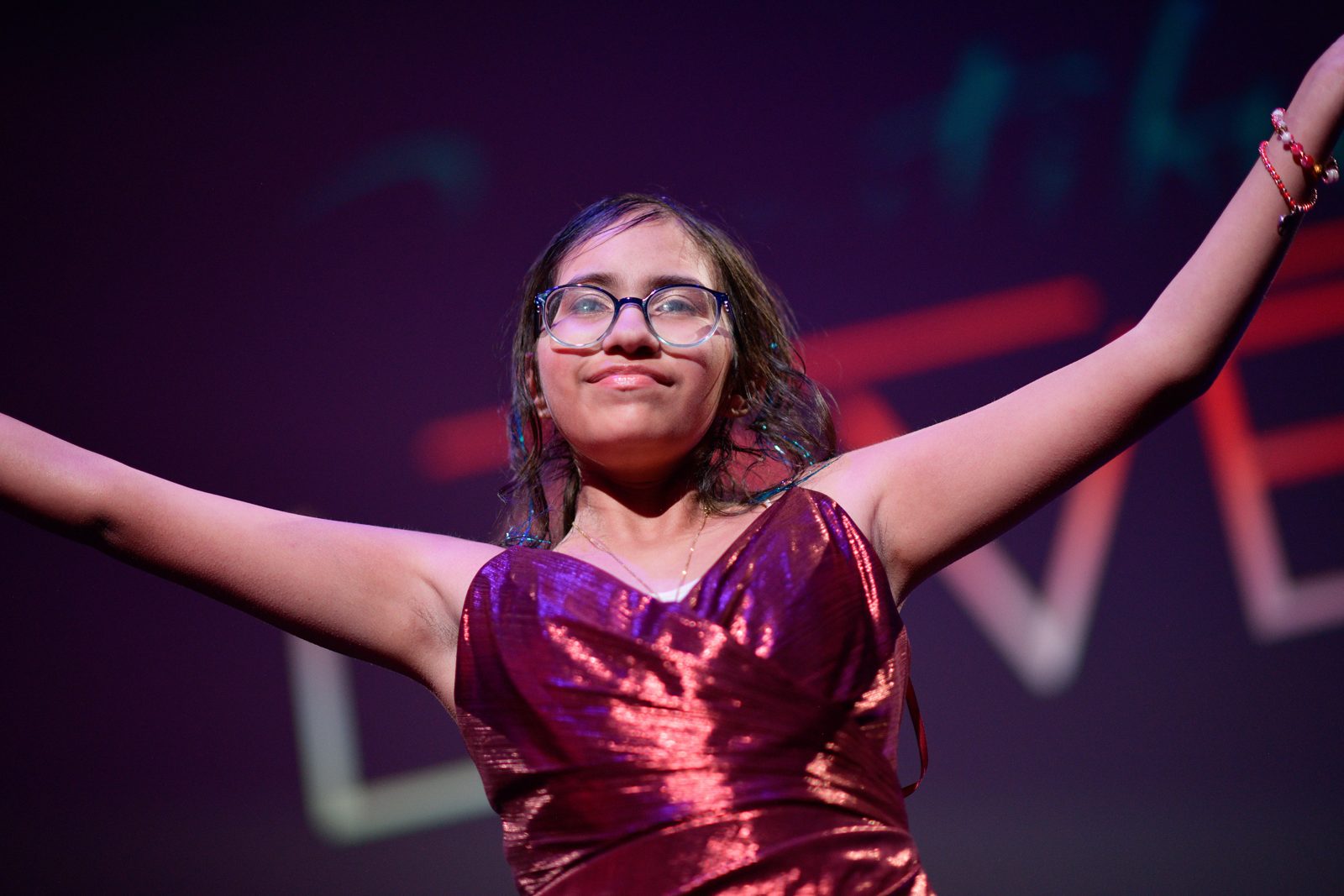 A young model in a shimmering red dress smiling with her arms raised on the fashion show stage