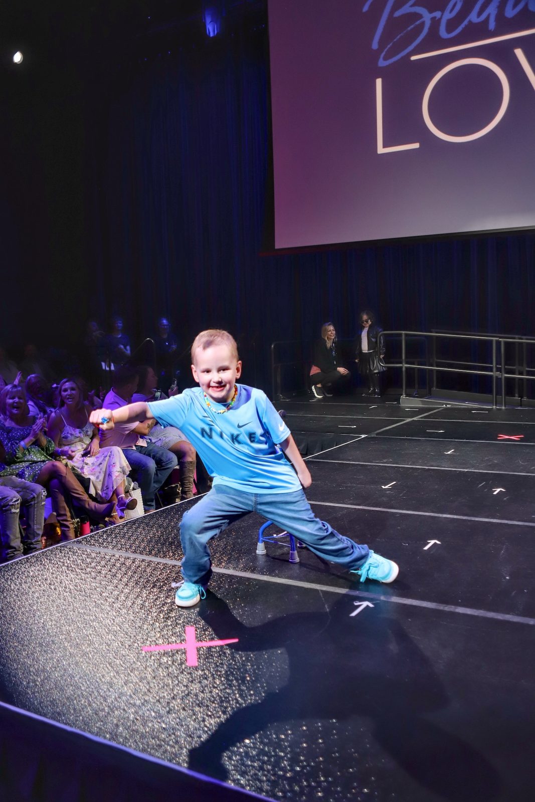 A boy in a blue tracksuit striking a dance pose on the fashion show runway